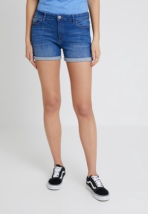 Persona vistiendo pantalones cortos de mezclilla azul enrollados, camiseta azul clara y zapatillas negras y blancas, de pie contra un fondo claro y sencillo.