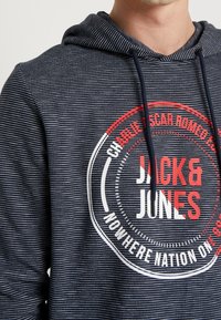 Felpa con cappuccio a righe navy con stringa di regolazione, caratterizzata da un logo circolare rosso e bianco che include il testo "JACK & JONES" e ulteriori dettagli.