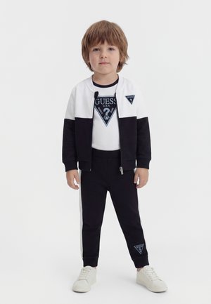 Ung gutt som står og har på seg en svart og hvit Guess-treningsdress med logoer på jakken, t-skjorten og buksene, samt hvite joggesko.