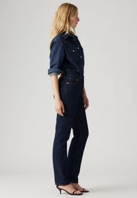 Donkerblauwe denimshirt met knoopsluiting aan de voorkant en twee borstzakken, gecombineerd met donkerblauwe rechtlopende jeans. Zwarte haksandalen maken de look compleet.
