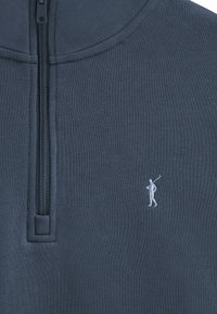 Halvdraget tröja i marinblått tyg. Har en texturerad yta och en liten vit golfarlogotyp. Dragkedjan sträcker sig upp till den höga kragen.