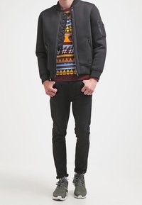 Giubbotto bomber nero con design trapuntato, maglione multicolore a fantasia sotto, pantaloni neri slim e sneakers grigie con suole bianche.