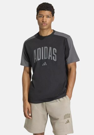 Junger Mann trägt ein schwarz-graues Adidas T-Shirt und beige Adidas-Shorts und steht mit einer Hand in der Tasche.
