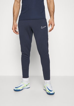 Navy Nike sweatpants with tapered legs and logo detail. Paired with grey and blue athletic shoes featuring neon accents. Cotton blend fabric.