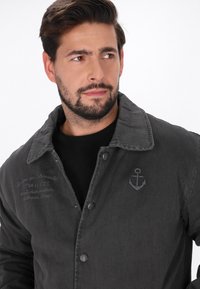 Donkergrijze denimjas met een knoopsluiting, grote kraag en subtiele ankerborduring op de borst. Kenmerkt zich door een gestructureerde stof.