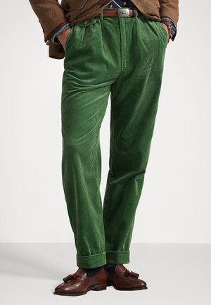 Zielone spodnie z korduroju o teksturowanym pionowym prążku, z plisowanym przodem i podwiniętymi mankietami, zestawione z brązowymi mokasynami.