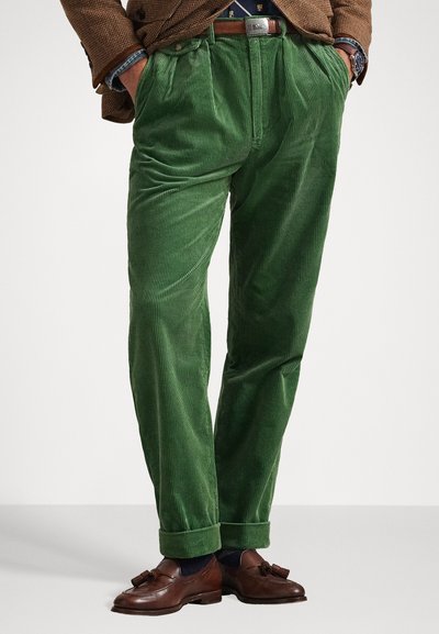 Pantaloni in corduroy verde con un motivo a coste verticali testurizzate, caratterizzati da una parte anteriore plissettata e risvolti, abbinati a mocassini marroni.