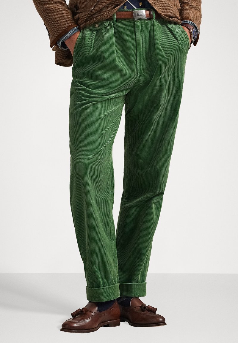 Zelene kordne hlače z teksturiranim navpičnim rebrastim vzorcem, ki imajo nameščen prednji del in zavihane hlačnice, v kombinaciji z rjavimi loaferji.
