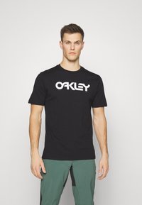 Sort bomulds t-shirt med korte ærmer, der har et stort hvidt "OAKLEY" logo på tværs af brystet. Rund halsudskæring.
