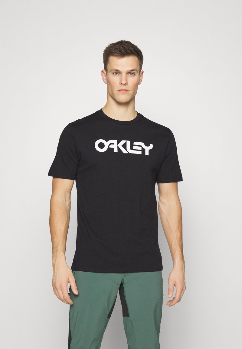 Sort bomulds t-shirt med korte ærmer, der har et stort hvidt "OAKLEY" logo på tværs af brystet. Rund halsudskæring.