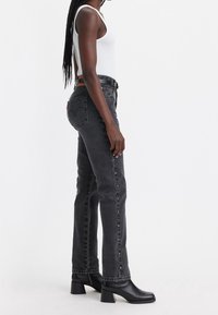 Mujer con cabello largo trenzado, vistiendo una camiseta sin mangas blanca, jeans negros de tiro alto con tachuelas en los lados, cinturón negro y botas de tobillo negras con tacón.