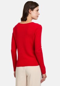 Pull en tricot rouge avec un col rond, des manches longues et des poignets côtelés. Le tissu semble doux et texturé, présentant une silhouette ajustée.