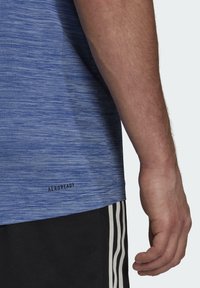 Siniset lyhythihaiset urheilupaidat, joissa on teksturoitu kuvio, mustalla "AEROREADY"-logolla helman lähellä, yhdistettynä mustiin shortseihin ja raidallisiin yksityiskohtiin.