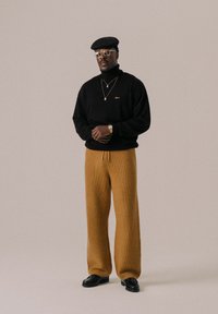Schwarzer gestrickter Rollkragenpullover, beige strukturierte weit geschnittene Hose, schwarze Schuhe und ein schwarzes Barett. Goldene Accessoires ergänzen das Outfit.