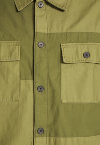 Camicia khaki con due tasche frontali, chiusura con bottoni e un design a blocchi di colore in diverse tonalità di verde. Tessuto di cotone testurizzato.