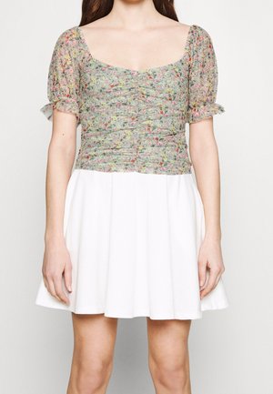 Woman wearing a floral ruched top with puffed sleeves and a square neckline paired with a white pleated skirt.