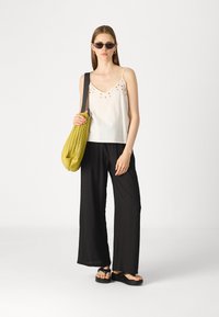 Haut sans manches blanc avec des détails brodés, pantalon noir à jambes larges, sac plissé jaune et sandales à plateforme noires.