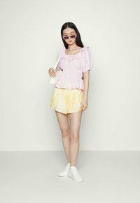 Blusa riscada em rosa e branco com mangas franzidas. Calções amarelos com efeito tie-dye. Ténis brancos e uma pequena mala branca. Óculos de sol escuros.