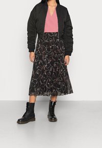 Schwarze Bomberjacke über einem rosa V-Ausschnitt-Top, kombiniert mit einem schwarzen Faltenrock mit einem weißen Linienzeichnungsmuster. Schwarze Schnürstiefel.