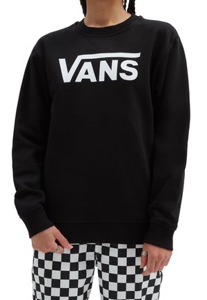 Henkilö käyttää mustaa Vans-hupparia, jossa on valkoinen logo, ja mustavalkoruudulliset housut, kädet rentoina sivuilla.