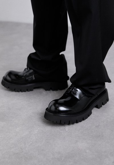Shiny black leather lace-up shoes with thick tread soles worn with long black pants on gray floor.