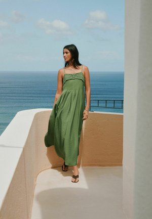 Frau in einem grünen ärmellosen Maxikleid, die auf einer beigen Terrasse steht und auf ein ruhiges blaues Meer unter einem teilweise bewölkten Himmel blickt.