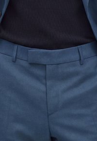 Gros plan sur un pantalon bleu sur mesure avec ceinture et passants, porté avec une chemise noire rentrée.
