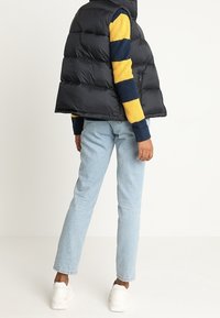 Czarna kamizelka puchowa z wysokim kołnierzem, noszona na niebiesko-żółtym paskowanym swetrze. Jasnoniebieskie dżinsy i białe trampki dopełniają stylizację.