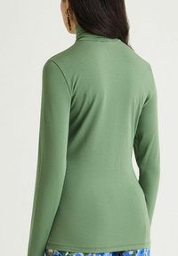 Maglia a collo alto a maniche lunghe in tessuto verde tenue, caratterizzata da un design aderente con texture liscia e cuciture minime.