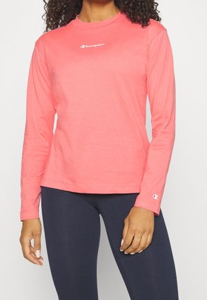 Woman wearing a coral long-sleeve Champion shirt and dark leggings, standing with arms relaxed by her sides.