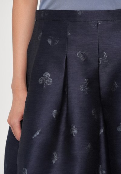 Dark navy pleated skirt with subtle faded playing card suit symbols printed, worn with a blue top, hand relaxed at side.