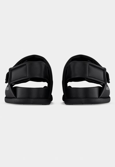 Μαύρα σανδάλια με σχεδιασμό δύο λουριών, ρυθμιζόμενους κλειστές Velcro και επίπεδη σόλα. Λείο υλικό και μινιμαλιστική αισθητική.