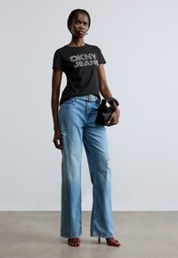 Svart T-skjorte med "DKNY JEANS" i sølvbokstaver, kombinert med lyseblå vidbente jeans med rift detalj, og røde høye hæler.