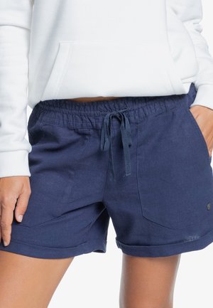 Person iført marineblå snor-shorts og en hvid langærmet sweatshirt med hænderne nær lommerne, beskåret ved taljen og lårene.