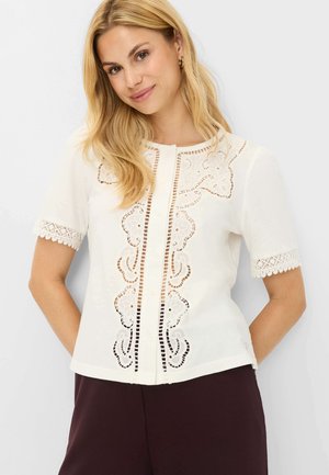 Blonde Frau trägt eine weiße Bluse mit kurzen Ärmeln, floraler Cut-Out-Stickerei und Spitzenbesatz, kombiniert mit dunkel gefärbter Hose.