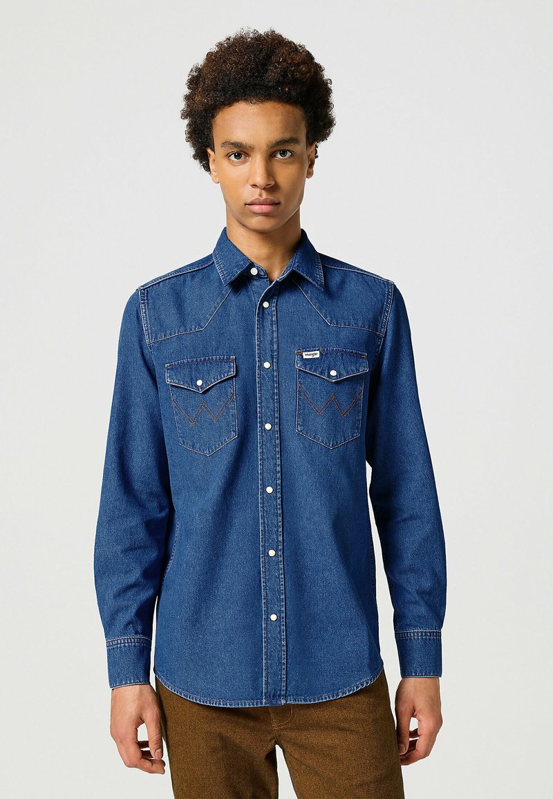 Camicia in denim a maniche lunghe di colore blu scuro, con due tasche sul petto, cuciture a contrasto, dettaglio del giogo appuntito e chiusura con bottoni.