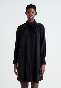 Robe noire à manches longues avec un nœud au cou, présentant une coupe droite, une fermeture à boutons sur le devant et des manches texturées. Tissu doux et lisse.
