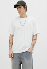 Hvid t-shirt med korte ærmer og en afslappet pasform, parret med lyseblå denimbukser. Et sølvkædelykke og en mørkegrøn kasket fuldender looket.