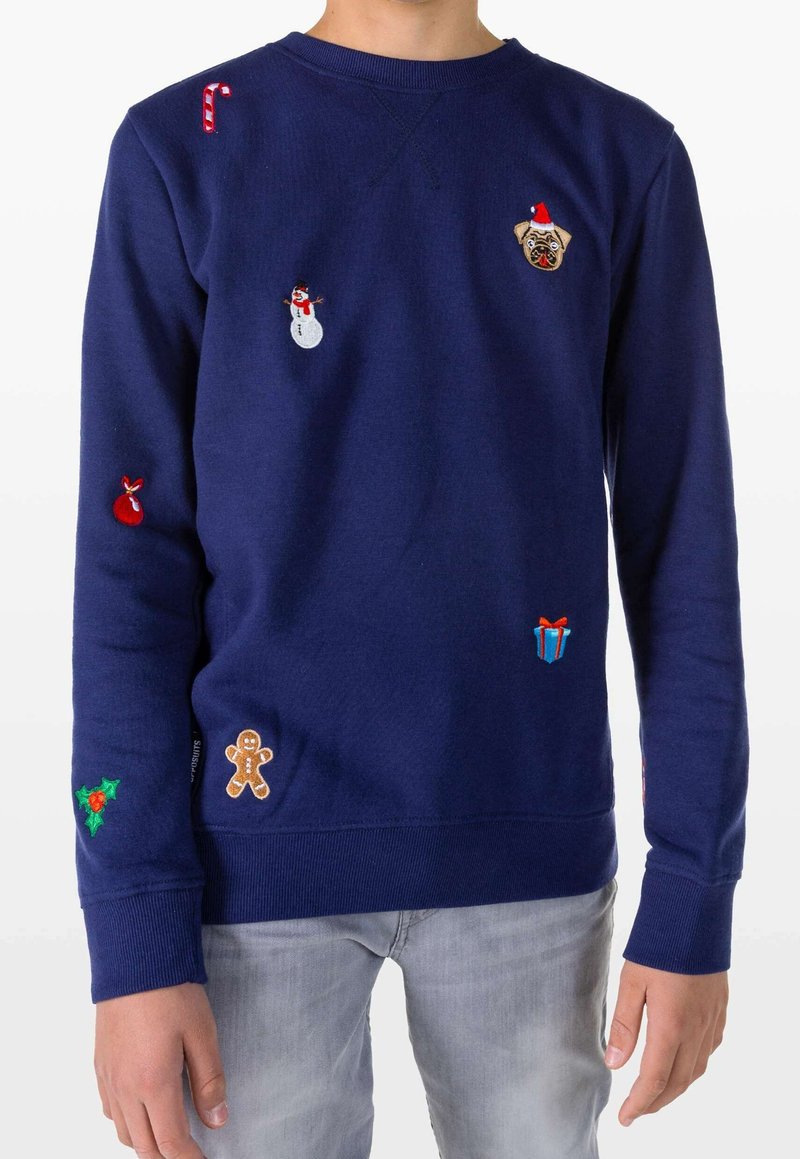 Pull bleu marine avec des patchs de Noël comprenant un sucre d'orge, un bonhomme de neige, un carlin portant un chapeau de Père Noël, un cadeau, du houx, un bonhomme en pain d'épice et une boule rouge.