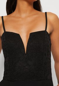 Cuerpo de encaje negro con un escote en V profundo, con tirantes finos y patrones florales, combinado con una tela negra lisa en la parte inferior.