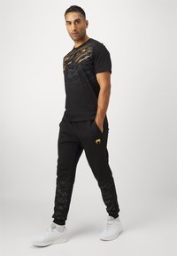 Musta urheiluvaatetus, jossa on teksturoidusta kankaasta valmistettu t-paita kultasilla yksityiskohdilla ja mustat verkkarit, joissa on hienovarainen kuvio. Valkoiset lenkkarit täydentävät asua.