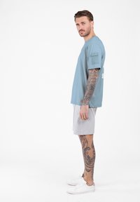Mann mit Ganzarm- und Bein-Tattoos, der ein blaues Kurzarmhemd, helle graue Shorts und weiße Turnschuhe trägt und im Profil steht.