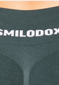 Ciemnozielony sportowy top na ramiączkach z prążkowanej tkaniny, szerokim dekoltem i białym logo "SMILODOX" na górnym pasie. Miękka, dopasowana faktura.