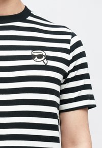 Czarnobiała koszulka w paski z okrągłym dekoltem. Na lewej piersi znajduje się mała naszywka z haftowaną kreskówkową twarzą w okularach przeciwsłonecznych.