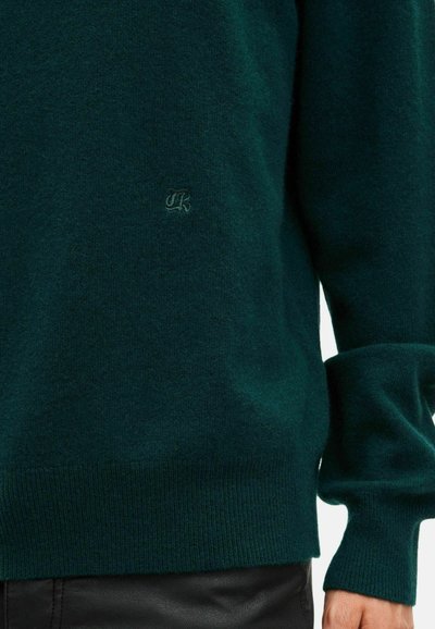 Ciemnozielony sweter dziany z małym haftowanym logo na piersi, noszony z czarnymi skórzanymi spodniami.