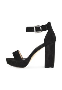 Sandalias de tacón en ante negro que cuentan con un grueso tacón cuadrado, correa para el tobillo con hebilla plateada y una amplia tira delantera. Planta interior suave en color beige.