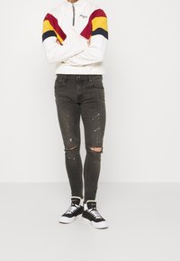 Suéter blanco con cremallera y rayas en rojo, amarillo y azul marino en las mangas, combinado con jeans negros rasgados con salpicaduras de pintura. Zapatillas negras.