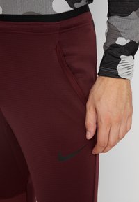 Nike sportbroeken in diep bordeauxrood, met textuurstof, zijzakken en een zwart logo op de linker bovenbeen.