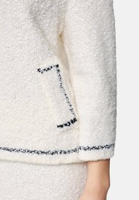 Weiße strukturierte Jacke mit schwarzem Besatz entlang der Taschenränder, Ärmelbündchen und Saum, getragen am Oberkörper und Arm einer Person.