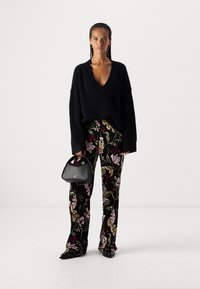 Schwarzer, übergroßer Strickpullover mit V-Ausschnitt, kombiniert mit schwarzen Hosen mit floralen Druck. Eine leichte Handtasche und elegante schwarze Schuhe runden den Look ab.